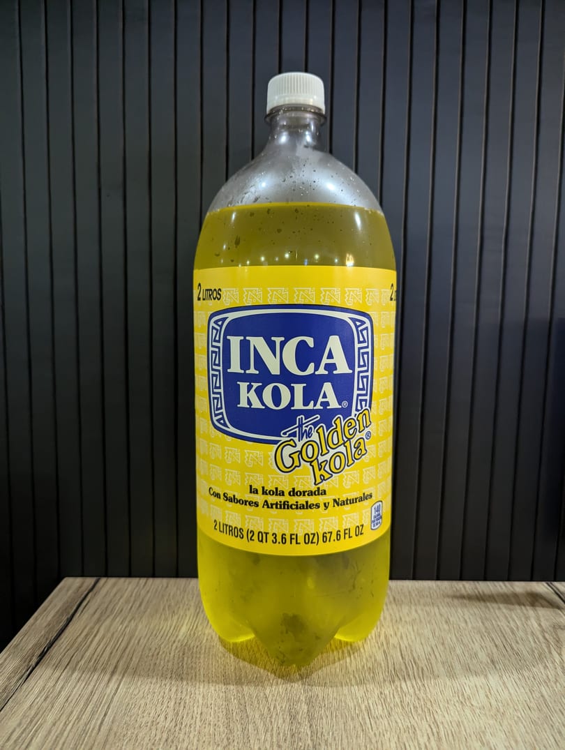 A 2-liter bottle of Inca Kola, "The Golden Kola," on a wooden surface with a dark background.