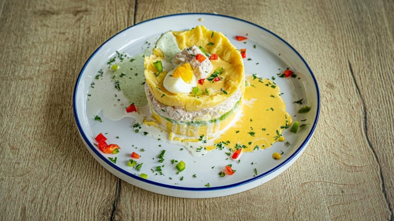Layered causa dish with mashed potatoes, tuna, avocado, egg, and colorful sauces on a white plate.