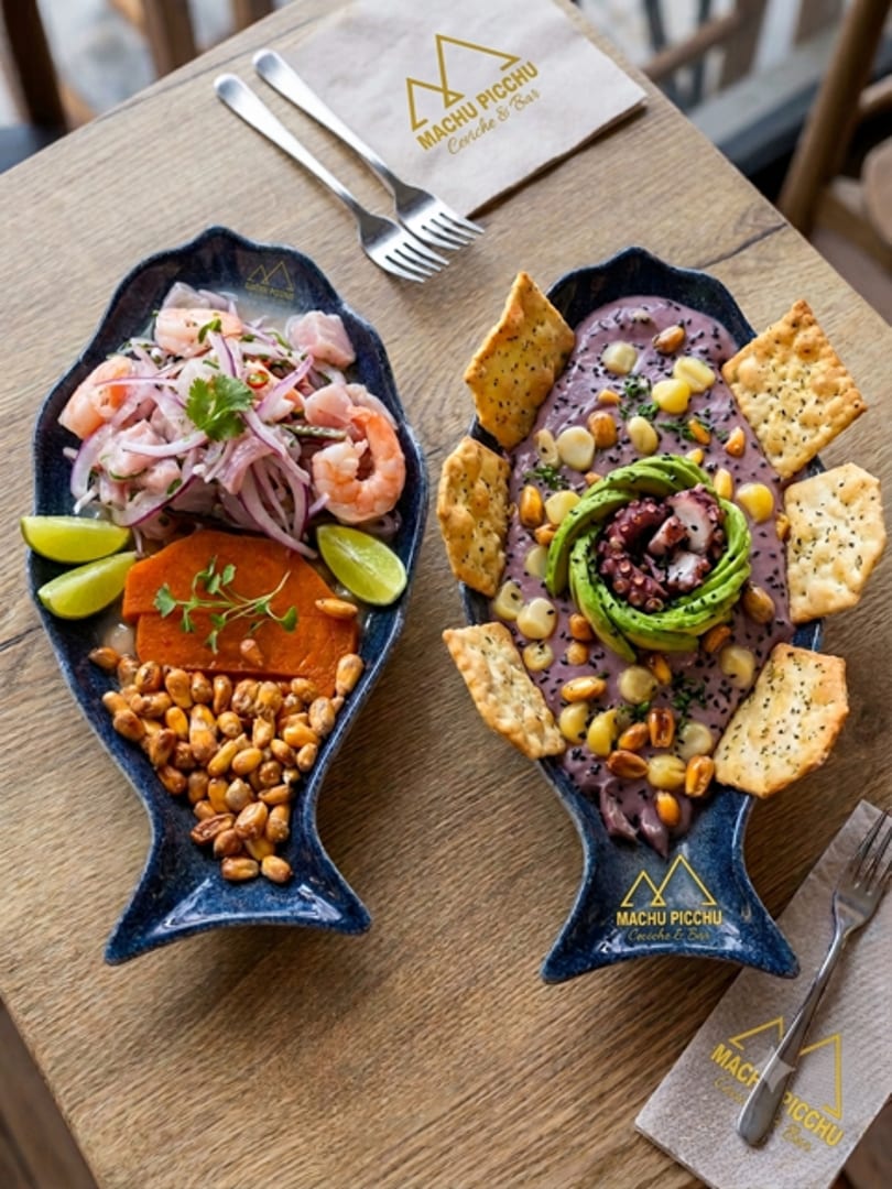 Two fish-shaped dishes of ceviche and octopus with avocado, crackers, and corn on a wooden table.