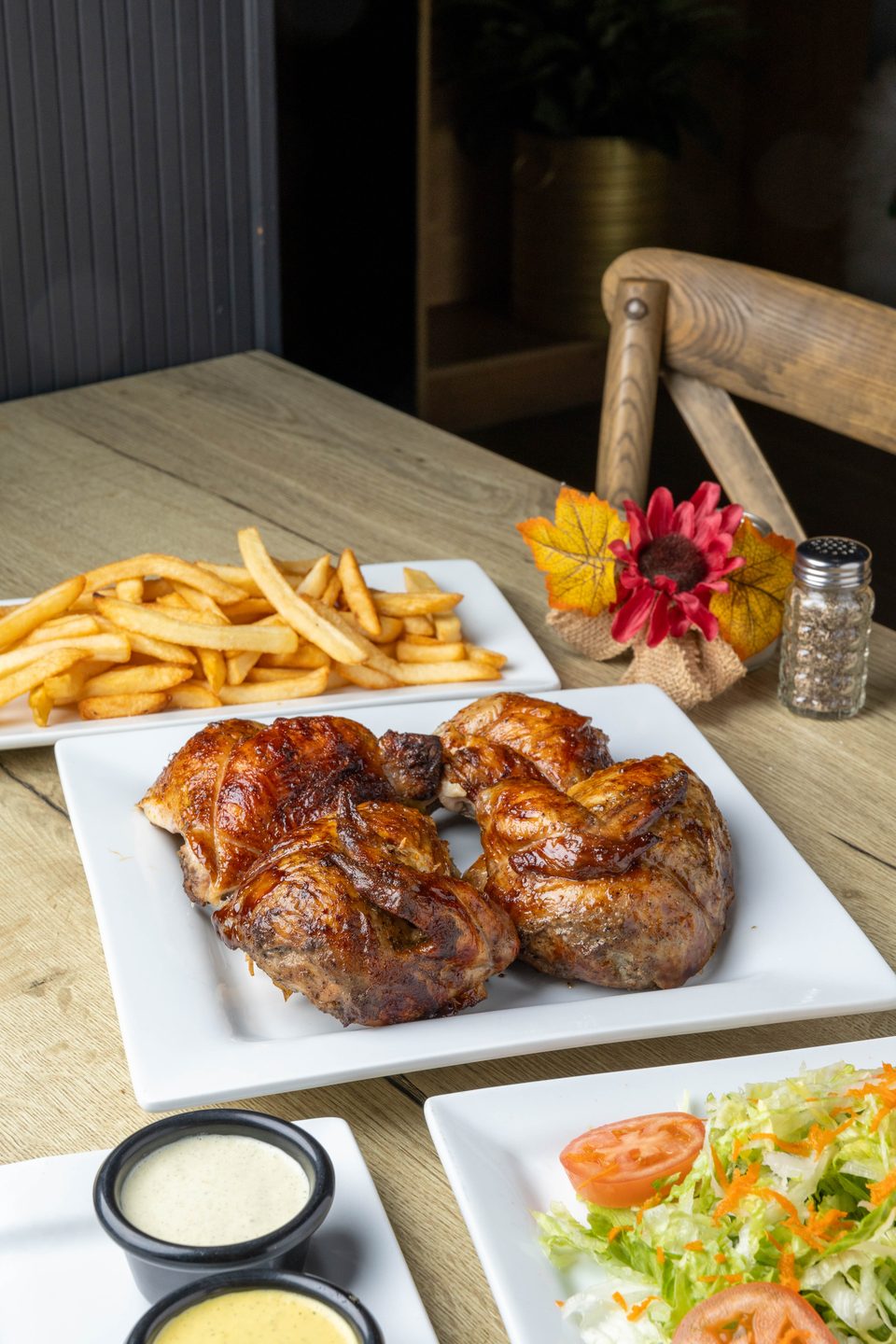 Plates of roasted chicken, french fries, and salad with dressing on a wooden table.