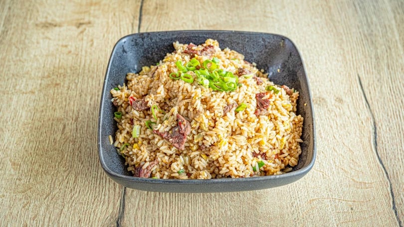 A black bowl filled with beef fried rice, garnished with chopped green onions, on a wooden table.