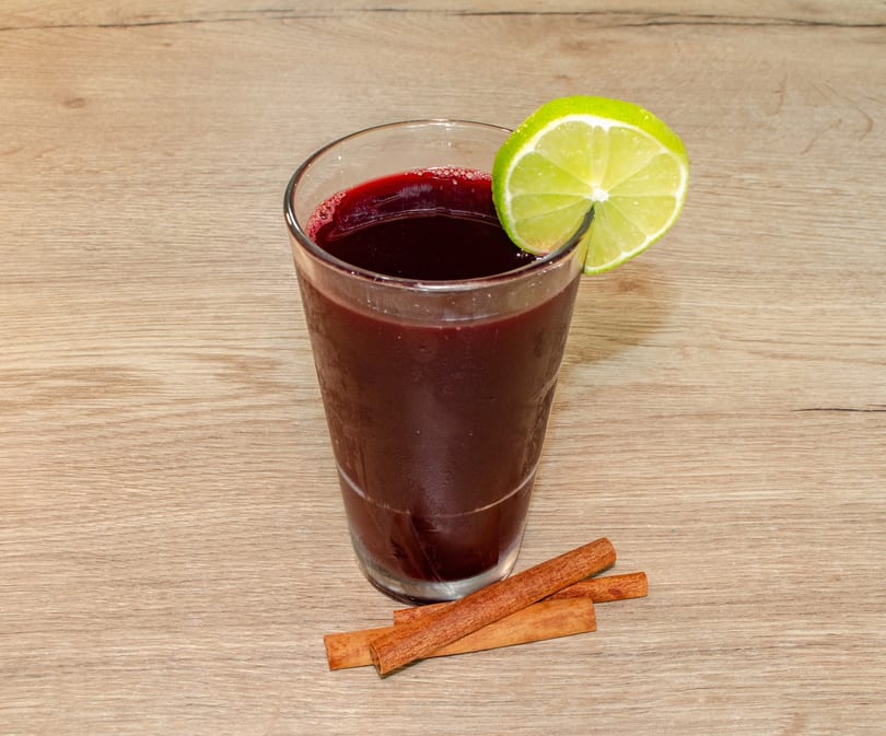 Dark red beverage in a glass, garnished with a lime slice and cinnamon sticks on a wooden surface.