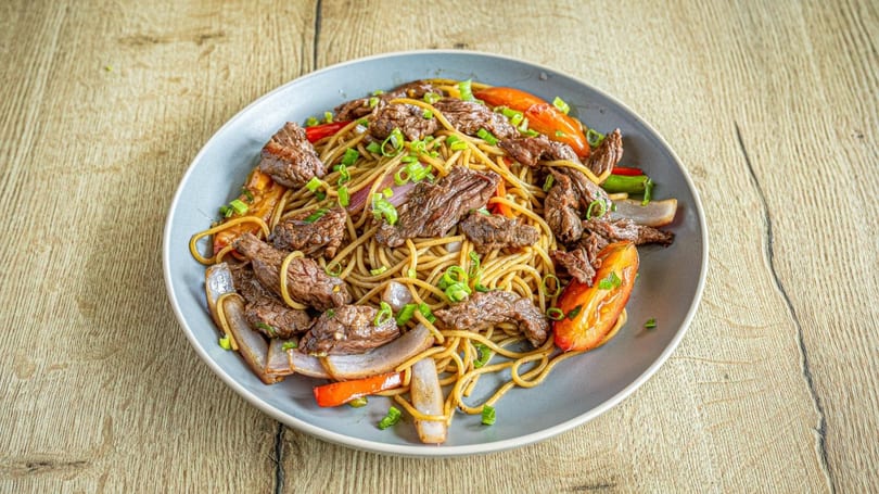 Lomo Saltado with noodles, beef, red onions, tomatoes, and green onions on a gray plate.