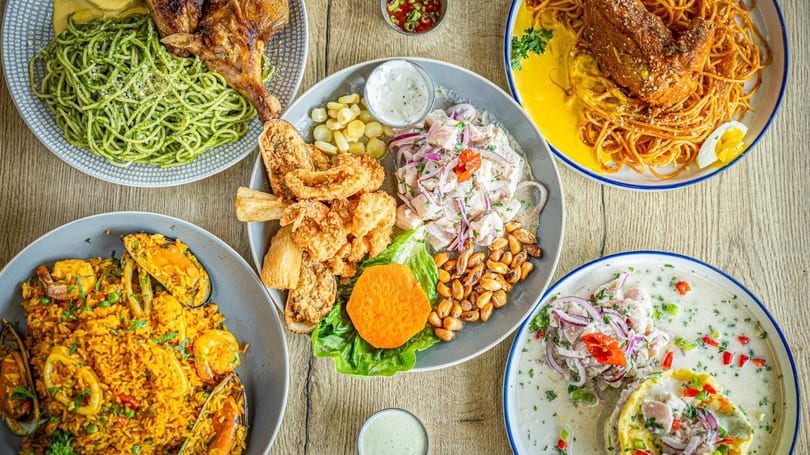 An overhead shot of a wooden table laden with various Peruvian dishes, including ceviche, fried seafood, and pasta.
